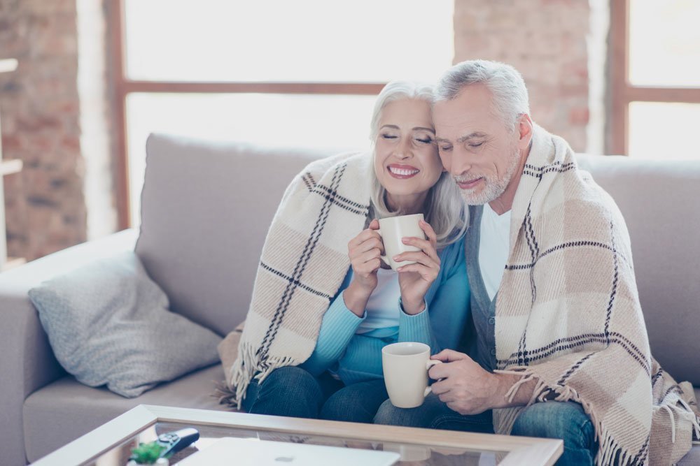 Älteres Paar mit einer Kuscheldecke und Tasse Tee auf dem Sofa sitzend