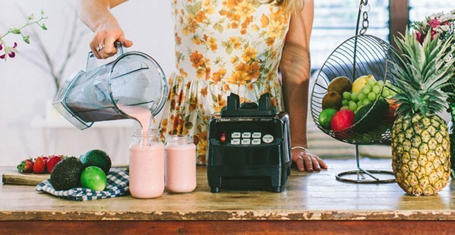 Frau im Kleid in einer Küche mit Obstkorb serviert rosa Smoothie aus dem Mixer in zwei Gläser